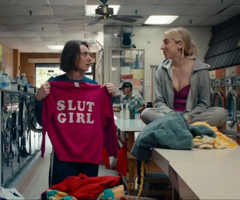 A still from the short film Sister!. In a laundromat, James (Julia Wendt), wearing a teal hockey jersey and dark pants, holds up a bright pink sweatshirt that reads "SLUT GIRL" in white letters, looking toward Tessa (Tessa Belle), who sits cross-legged on a folding table surrounded by laundry, smiling back. Washing machines line both walls in the background.