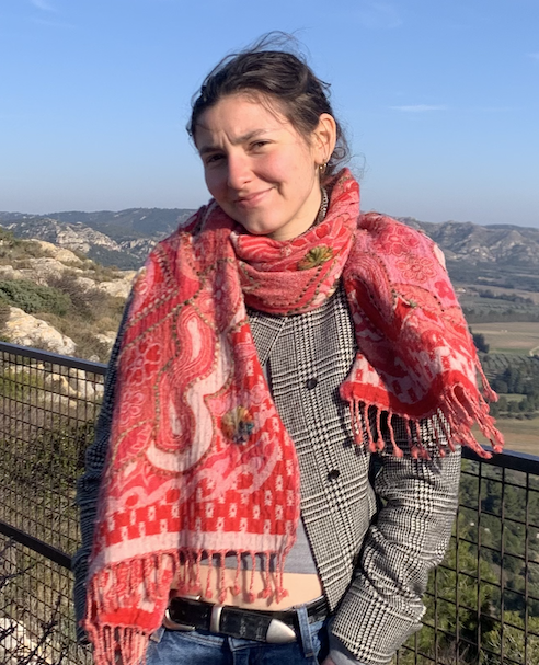 "I Promise It's Possible": Hannah Richman’s Story A young woman stands on an outdoor overlook with a metal railing, smiling softly at the camera. She wears a vibrant red paisley fringed scarf over a gray plaid blazer with jeans and a black belt. Behind her stretches a scenic landscape of rolling hills, rocky outcroppings, green fields, and cypress trees under a clear blue sky.