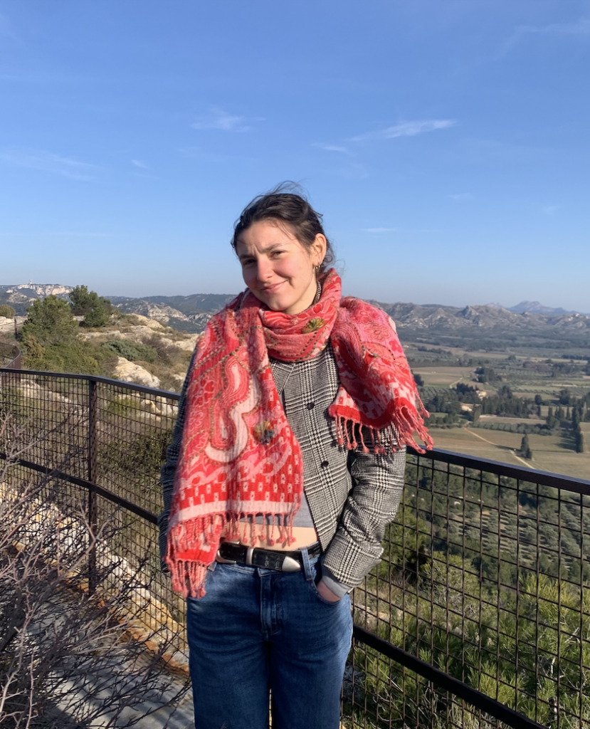 A young woman stands on an outdoor overlook with a metal railing, smiling softly at the camera. She wears a vibrant red paisley fringed scarf over a gray plaid blazer with jeans and a black belt. Behind her stretches a scenic landscape of rolling hills, rocky outcroppings, green fields, and cypress trees under a clear blue sky.
